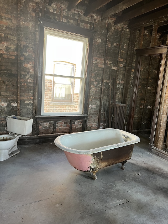 bathroom featuring beamed ceiling, a bath, toilet, and brick wall