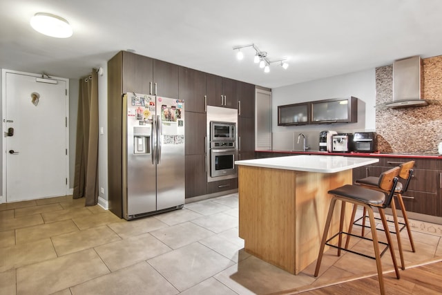 kitchen featuring a breakfast bar, backsplash, wall chimney range hood, appliances with stainless steel finishes, and dark brown cabinetry