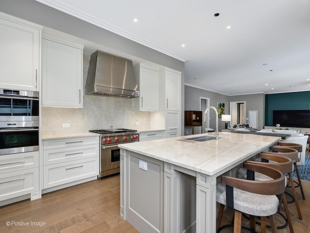 kitchen with sink, appliances with stainless steel finishes, white cabinetry, a center island with sink, and wall chimney exhaust hood