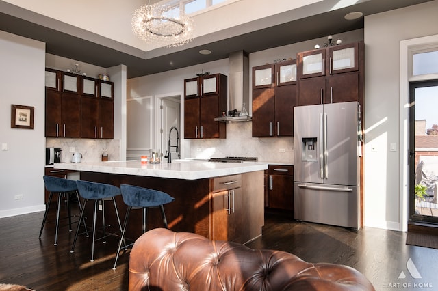 kitchen featuring wall chimney exhaust hood, stainless steel fridge, decorative backsplash, an island with sink, and decorative light fixtures