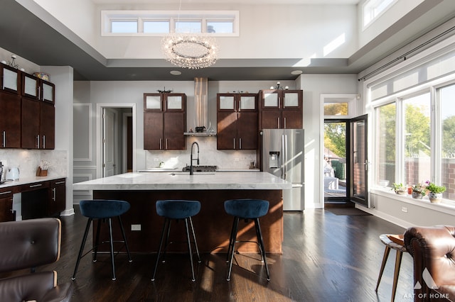 kitchen featuring an inviting chandelier, stainless steel refrigerator with ice dispenser, wall chimney exhaust hood, an island with sink, and dark brown cabinetry