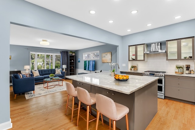 kitchen with double oven range, a kitchen island with sink, wall chimney exhaust hood, light hardwood / wood-style floors, and a kitchen bar