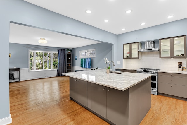 kitchen featuring wall chimney exhaust hood, a kitchen island with sink, sink, light hardwood / wood-style floors, and stainless steel range with gas cooktop