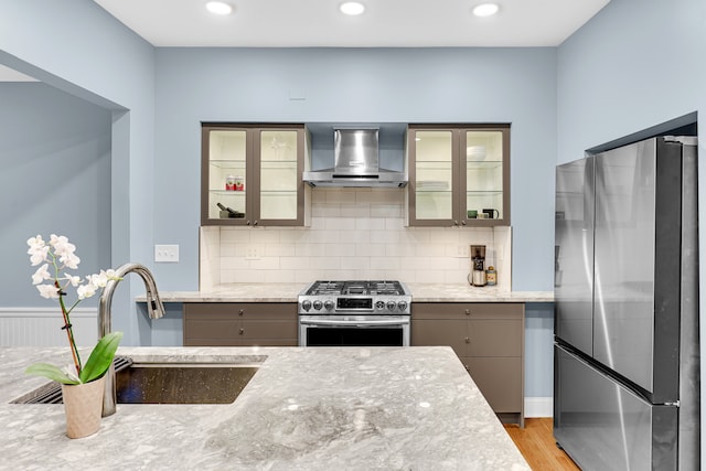 kitchen with wall chimney exhaust hood, light hardwood / wood-style floors, light stone countertops, and appliances with stainless steel finishes