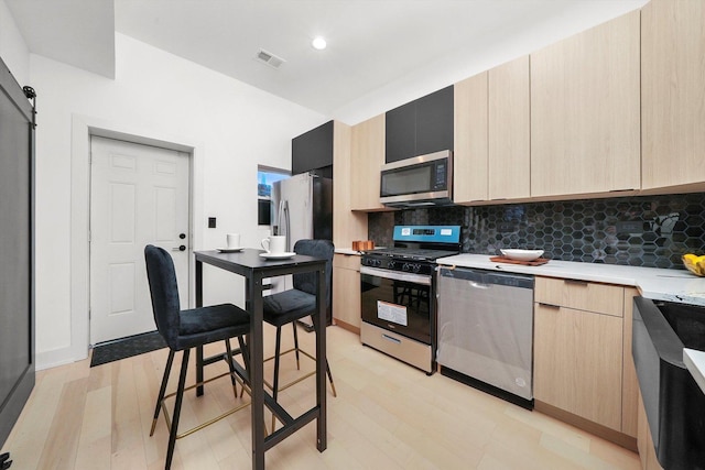 kitchen with a barn door, tasteful backsplash, light wood-type flooring, light brown cabinetry, and appliances with stainless steel finishes