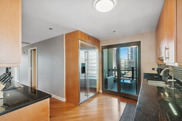 kitchen featuring sink, light hardwood / wood-style flooring, dark stone countertops, decorative backsplash, and stainless steel built in fridge