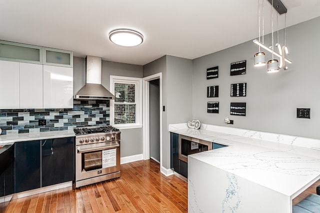 kitchen featuring decorative backsplash, appliances with stainless steel finishes, wall chimney exhaust hood, decorative light fixtures, and light hardwood / wood-style flooring