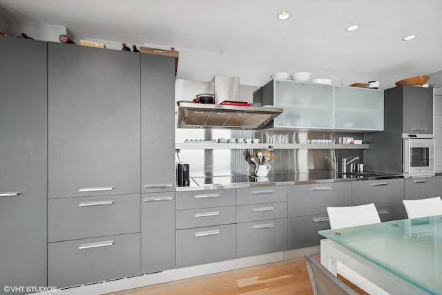 kitchen featuring wall chimney exhaust hood, stainless steel oven, stainless steel counters, sink, and light hardwood / wood-style flooring