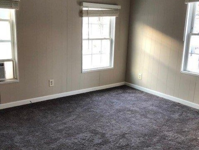 unfurnished room with wooden walls, plenty of natural light, baseboards, and dark colored carpet