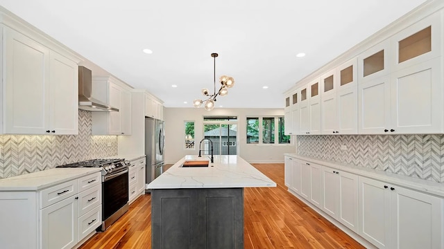 kitchen featuring wall chimney exhaust hood, stainless steel appliances, sink, pendant lighting, and an island with sink