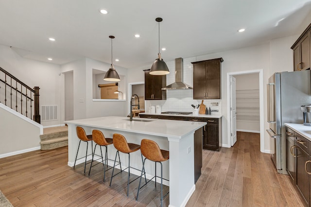 kitchen featuring appliances with stainless steel finishes, light wood-type flooring, wall chimney exhaust hood, a kitchen island with sink, and sink