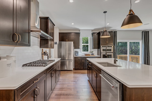 kitchen featuring wall chimney exhaust hood, an island with sink, pendant lighting, appliances with stainless steel finishes, and light wood-type flooring