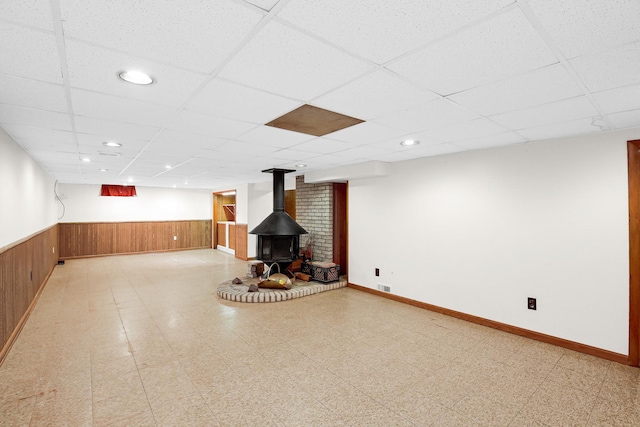 basement with a wood stove, wooden walls, and a drop ceiling