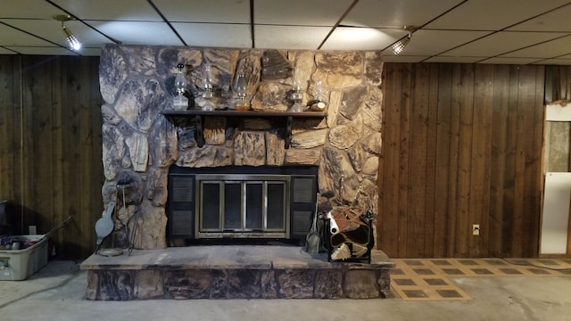 interior details featuring concrete floors, a stone fireplace, a drop ceiling, and wooden walls