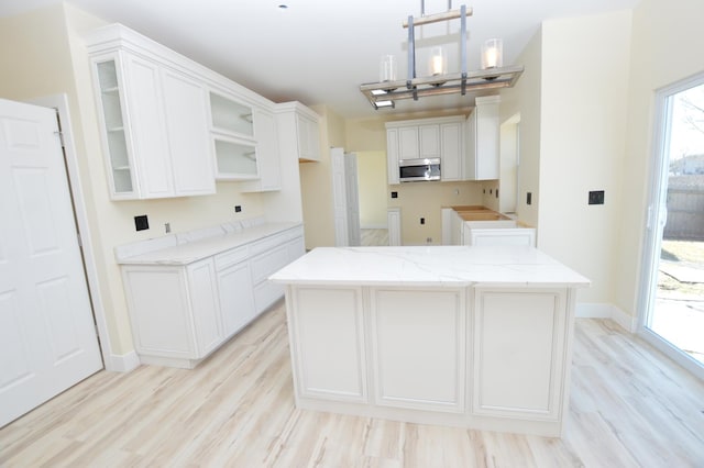 kitchen with stainless steel microwave, a kitchen island, glass insert cabinets, light wood-style floors, and white cabinetry