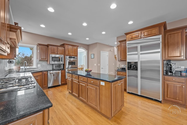 kitchen with sink, built in appliances, dark stone countertops, light wood-type flooring, and a kitchen island