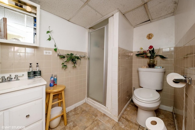 bathroom with a paneled ceiling, vanity, toilet, tile walls, and an enclosed shower