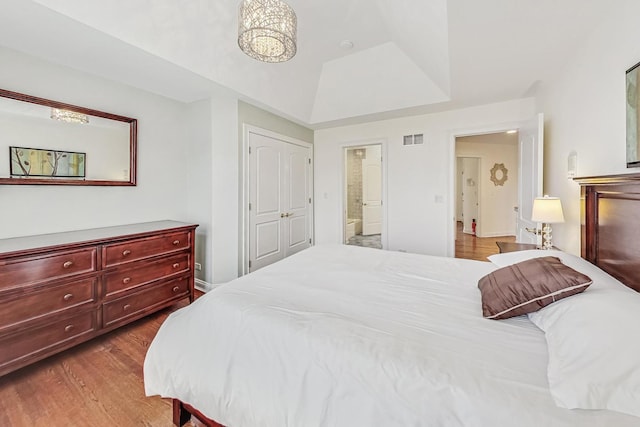 bedroom with ensuite bath, wood finished floors, visible vents, vaulted ceiling, and a raised ceiling