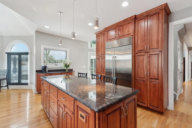 kitchen with dark stone countertops, hanging light fixtures, a center island, built in fridge, and light wood-type flooring