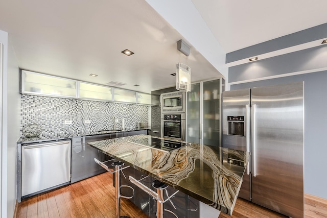 kitchen featuring wood-type flooring, decorative light fixtures, appliances with stainless steel finishes, dark stone counters, and decorative backsplash