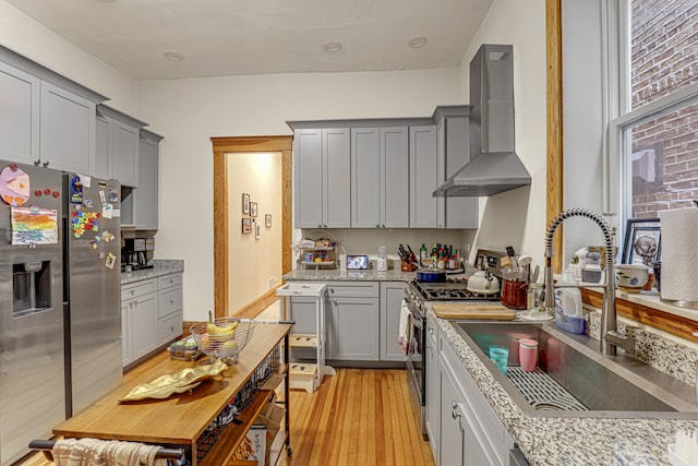 kitchen featuring light stone countertops, sink, wall chimney range hood, light hardwood / wood-style floors, and appliances with stainless steel finishes