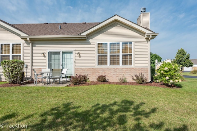 rear view of house featuring a patio, brick siding, a lawn, roof with shingles, and a chimney