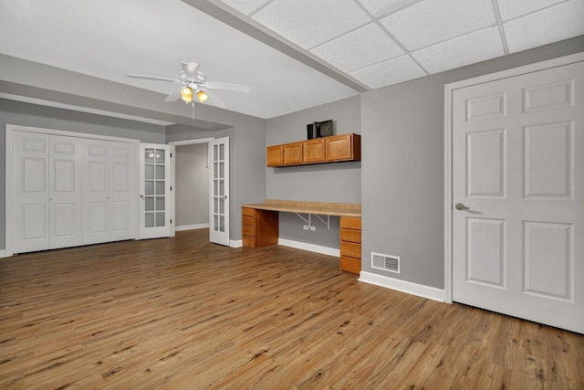 unfurnished living room featuring french doors, light wood-type flooring, a paneled ceiling, ceiling fan, and built in desk