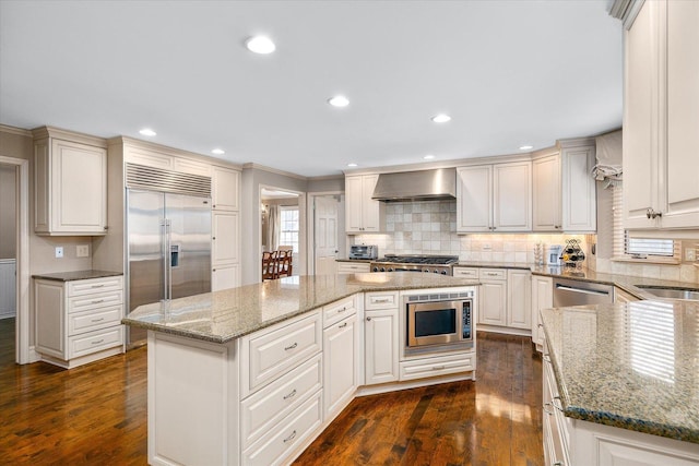 kitchen with wall chimney exhaust hood, a kitchen island, dark wood-style flooring, built in appliances, and light stone countertops