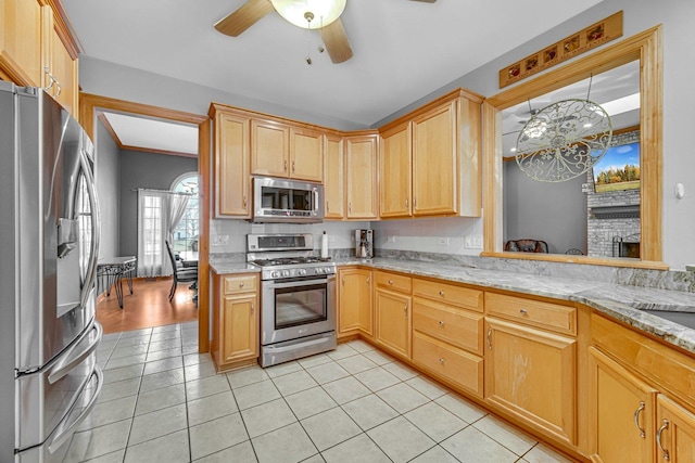 kitchen with light stone countertops, stainless steel appliances, a brick fireplace, light tile patterned flooring, and ceiling fan with notable chandelier