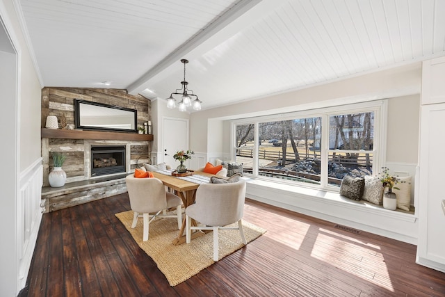 dining area featuring vaulted ceiling with beams, a chandelier, a stone fireplace, wainscoting, and dark wood-style floors