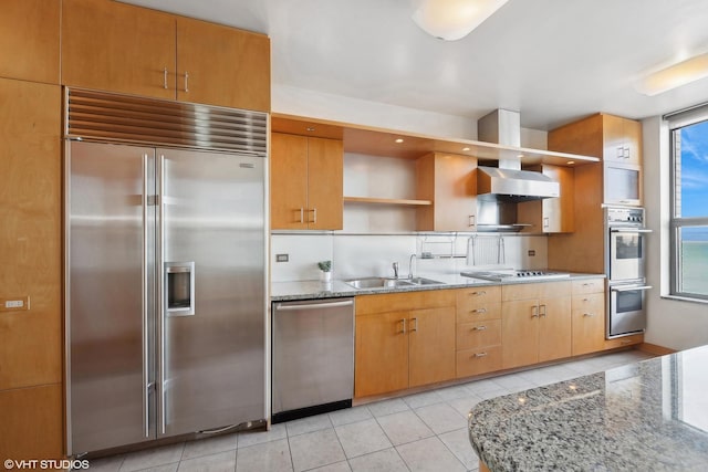 kitchen with light stone counters, stainless steel appliances, wall chimney range hood, sink, and light tile patterned floors