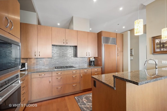 kitchen with light wood-type flooring, built in appliances, dark stone countertops, a center island, and hanging light fixtures