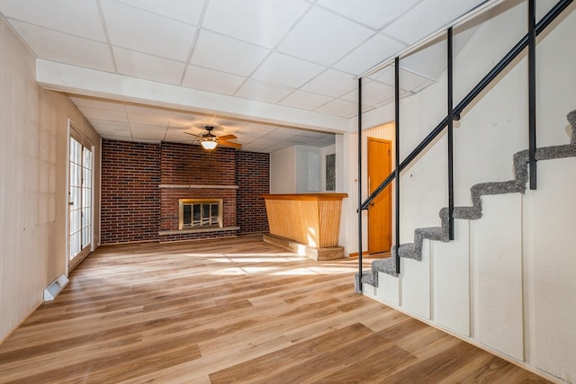 unfurnished living room with a paneled ceiling, ceiling fan, wood-type flooring, brick wall, and a brick fireplace