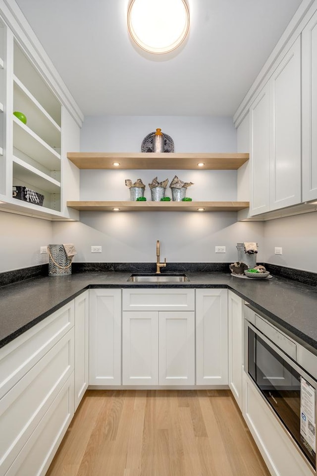 kitchen with a sink, stainless steel microwave, open shelves, dark countertops, and light wood finished floors