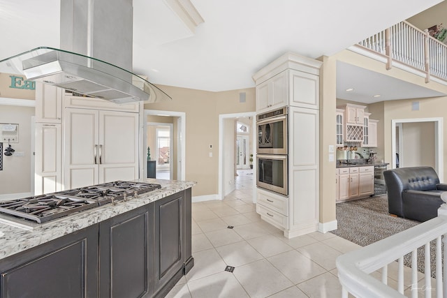 kitchen with cream cabinetry, light tile patterned flooring, light stone counters, island exhaust hood, and stainless steel appliances