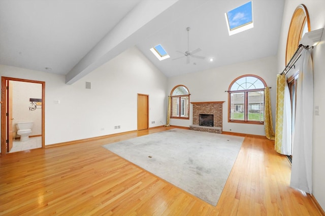 unfurnished living room with a skylight, ceiling fan, a brick fireplace, high vaulted ceiling, and light wood-type flooring