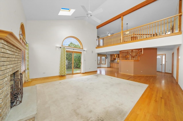 unfurnished living room with a skylight, ceiling fan, a brick fireplace, high vaulted ceiling, and light hardwood / wood-style floors