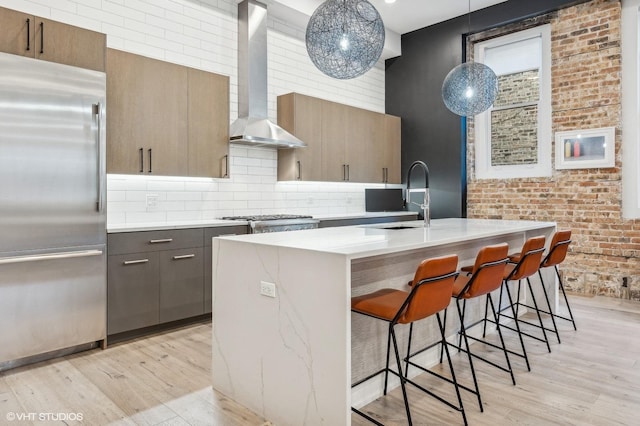 kitchen with appliances with stainless steel finishes, wall chimney exhaust hood, a kitchen island with sink, pendant lighting, and light hardwood / wood-style floors