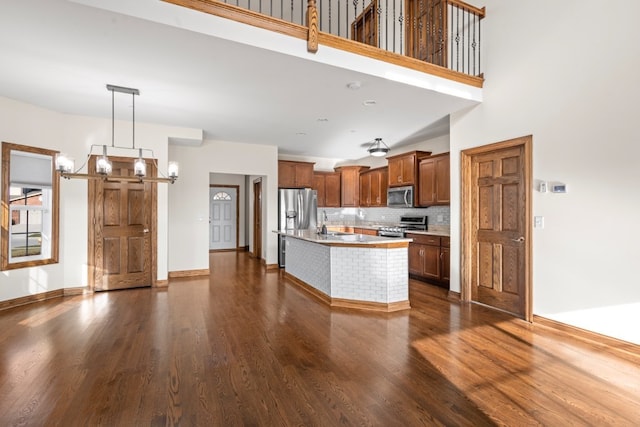 kitchen featuring a towering ceiling, an island with sink, decorative light fixtures, dark hardwood / wood-style flooring, and stainless steel appliances