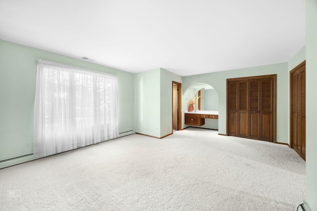 unfurnished bedroom with baseboard heating, light colored carpet, and two closets