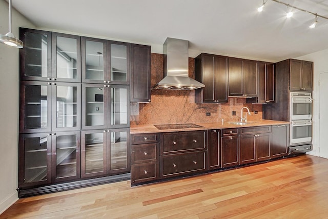 kitchen with black electric stovetop, wall chimney exhaust hood, stainless steel double oven, sink, and light hardwood / wood-style flooring