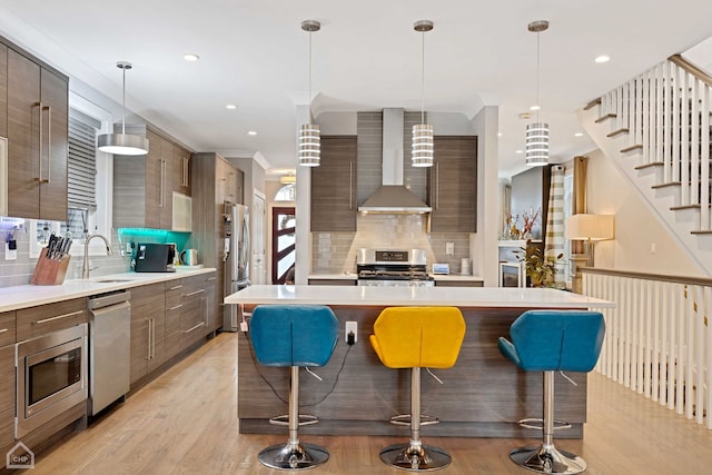 kitchen with appliances with stainless steel finishes, pendant lighting, a kitchen bar, light wood-type flooring, and wall chimney exhaust hood