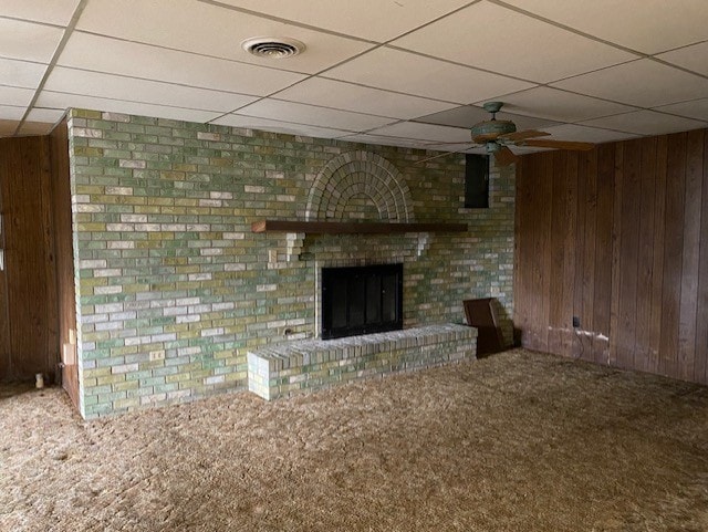 unfurnished living room with carpet, a drop ceiling, wooden walls, a brick fireplace, and ceiling fan