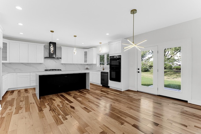 kitchen with wall chimney exhaust hood, hanging light fixtures, light hardwood / wood-style floors, white cabinets, and black appliances