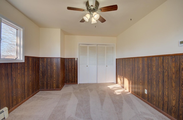 unfurnished bedroom featuring ceiling fan, baseboard heating, wood walls, light colored carpet, and a closet