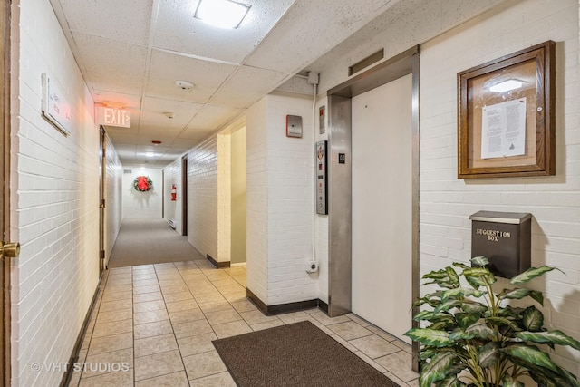 hallway with brick wall, a drop ceiling, light tile patterned flooring, and elevator