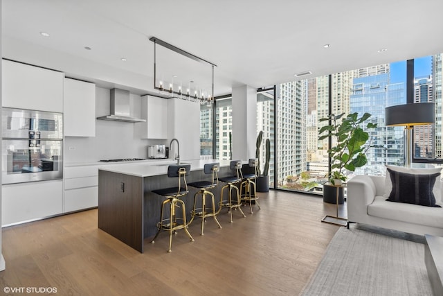 kitchen with wall chimney exhaust hood, gas stovetop, a kitchen island with sink, light hardwood / wood-style floors, and white cabinetry