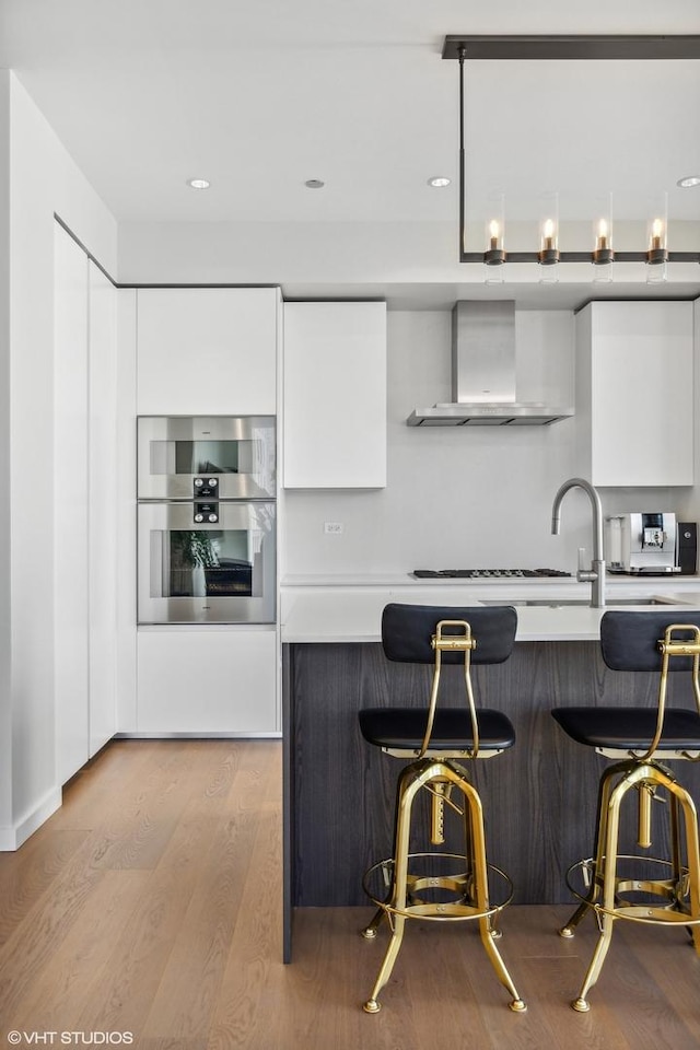 kitchen featuring wall chimney exhaust hood, a breakfast bar, stainless steel double oven, light hardwood / wood-style flooring, and white cabinetry