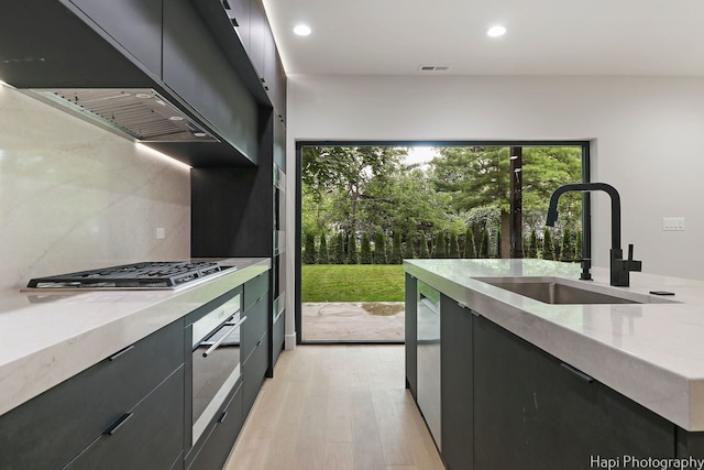 kitchen featuring decorative backsplash, light wood-type flooring, wall chimney exhaust hood, stainless steel appliances, and sink