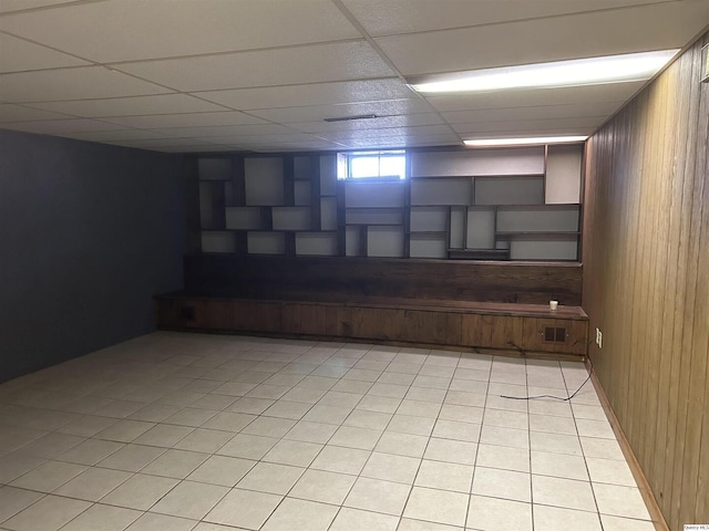 basement featuring light tile patterned floors, a drop ceiling, and wooden walls